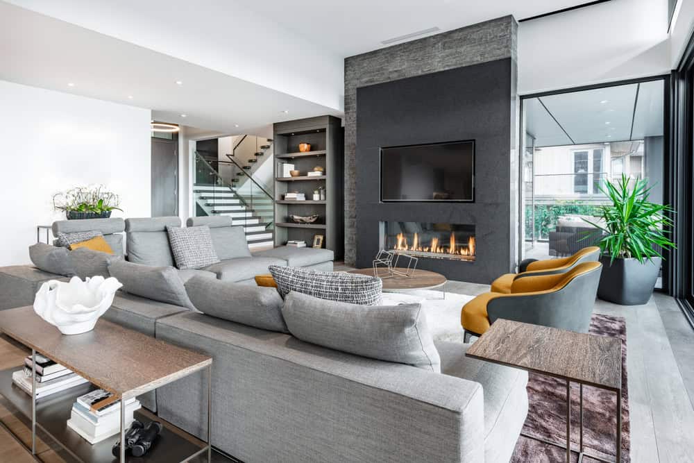 A modern living room featuring a large gray sectional sofa, two mustard yellow armchairs, and a sleek black coffee table. The focal point is a contemporary fireplace set in a dark stone wall with a mounted TV above, while large windows and strategically placed mirrors provide ample natural light.