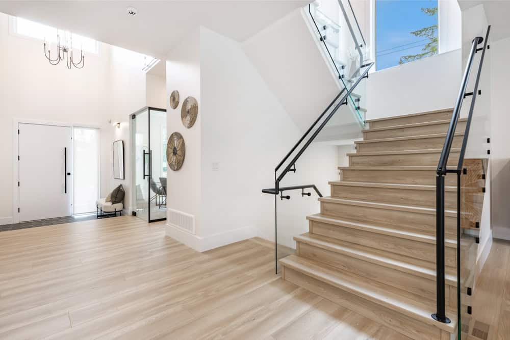 A spacious and bright entryway featuring light wood flooring. To the right, a wooden staircase with black railings leads up. The white walls are adorned with decorative mirrors from Litchfield County. An open door reveals a glimpse into another room with a chair and a chandelier hanging above.