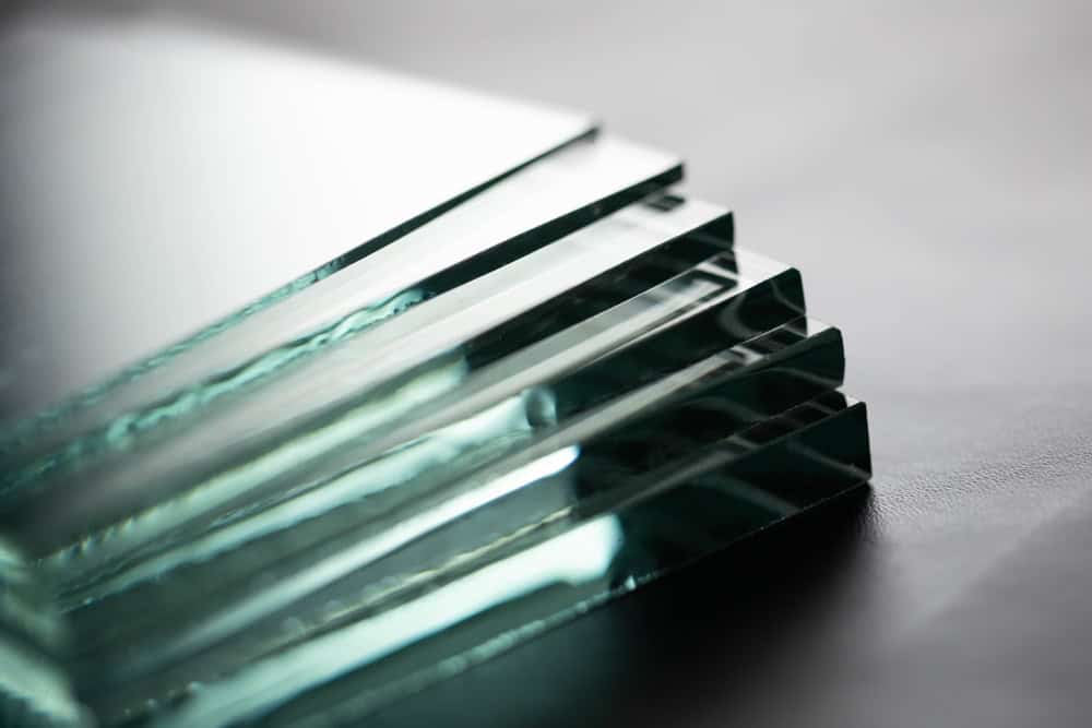 Close-up image of several sheets of glass stacked on top of each other in a fanned-out arrangement, showing their smooth edges and clear, reflective surfaces. The background is an out-of-focus dark gray, highlighting the clarity of the glass.